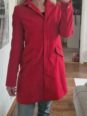 Röd klassisk kappa med krage - Stilren röd kappa med klassisk krage och två fickor framtill med lock. Kappan har dold knäppning och raka ärmar. Perfekt för dig som vill sticka ut med färg i vår eller på hösten..