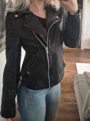 Svart biker skinnjacka med dragkedjor - Snygg svart skinnjacka i bikerstil med silvriga dragkedjor och detaljer. Jackan har quiltade partier på axlar och ärmar samt snedställd dragkedja framtill. Två fickor med dragkedja och justerbara spännen i sidorna. Perfekt för en cool och trendig look. Äkta skinn. Lite för stor för mig, har i vanliga fall XS och jackan är storlek M.