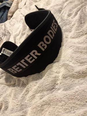 Svart lyftarbälte Better Bodies - Snyggt svart lyftarbälte från Better Bodies med tydlig logga i grått. Bältet är brett och vadderat för extra stöd vid tunga lyft och har kardborrestängning för enkel justering. Perfekt för gymmet och styrketräning.