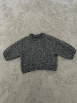 Grå stickad tröja från Gina Tricot - Mysig grå stickad tröja från Gina Tricot i storlek XS. Tröjan har en avslappnad passform, ribbade muddar vid hals, ärmslut och nederkant samt långa ärmar. Perfekt för lager på lager och enkel att matcha med jeans eller kjol.