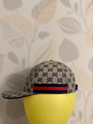 Gucci keps med monogram och röd-blå rand - Snygg keps från Gucci i beige med mörkblått GG-monogram över hela kepsen. Klassisk böjd skärm och en ikonisk röd-blå rand på sidan. Tillverkad i canvasmaterial som ger en exklusiv känsla och streetstyle-vibe.