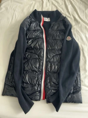 Svart pufferjacka från Moncler - Snygg svart pufferjacka från Moncler med quiltad front och rygg i glansigt material samt stickade ärmar och sidor. Dragkedja framtill med röd, vit och blå kant. Moncler-logga på vänster ärm. Perfekt för kalla vinterdagar. Inte äkta.