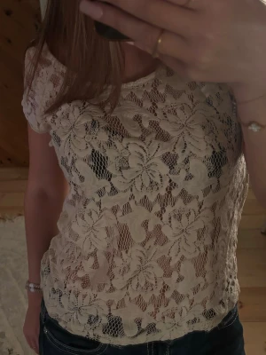 Beige spetsblus med blommönster - Säljer en superfin beige blus i spets med stora blommiga mönster. Blusen är lätt transparent, har korta ärmar och rund halsringning. Perfekt att styla med ett linne under och ett par jeans för en fräsch look.