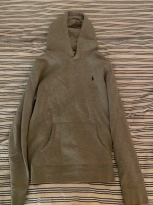 Grå hoodie från Polo Ralph Lauren - Snygg grå hoodie från Polo Ralph Lauren med klassisk huva och känguruficka framtill. Diskret svart logga broderad på bröstet. Tillverkad i mjukt bomullsmaterial som är perfekt för en avslappnad stil.