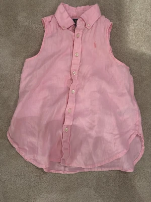 Rosa ärmlös linneskjorta Polo Ralph Lauren -  Snygg rosa ärmlös skjorta från Polo Ralph Lauren i luftigt linne. Klassisk krage, knappar framtill och broderad logga på bröstet i samma färg. Perfekt för varma dagar och enkel att styla till olika looks. Storlek är 12 men är liten i storlek.