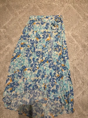 Blommig omlottkjol i blått, orange, vitt och gult - Säljer en lång omlottkjol med blommigt mönster i vitt, gul, orange och lite olika nyanser av blå. Kjolen har knytband i midjan och ett resårband baktill vilket gör att den blir stretchig i midjan och sitter inte åt. Är gjord i ett lätt och luftigt material, perfekt för varma dagar. Passar dig som gillar färgglada och somriga plagg.