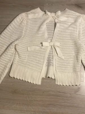 Vit stickad kofta med rosetter - Fin vit stickad kofta med två rosetter framtill. Koftan har långa ärmar. Materialet känns mjukt och bekvämt. 