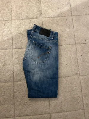 Blå jeans från Dondup - Säljer ett par klassiska blå jeans från Dondup med snygg tvätt och gul kontrastsöm. Jeansen har en bakficka med dekorativ detalj och ett svart läderpatch med logga i midjan. Perfekta för en avslappnad och trendig look.