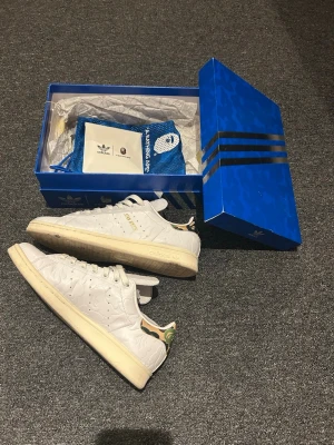 Adidas x BAPE Stan Smith vita sneakers - Unika vita Adidas x BAPE Stan Smith. Skorna har präglat mönster med BAPE:s ikoniska kamouflage och guldiga detaljer på plösen. Klassisk låg modell med vit sula och gröna detaljer på hälen. Levereras i originalkartong med extra påse och extra BAPE camo skosnören.          Priset går att diskutera vid snabb affär!
