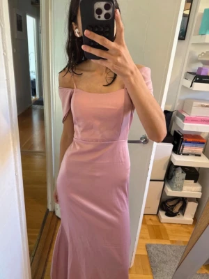 Wall g balklänning - Säljer en elegant ljusrosa långklänning med offshoulder och smala axelband. Klänningen har figurnära passform och en stilren design som ger en romantisk känsla. Perfekt för speciella tillfällen.