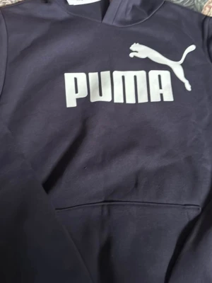 Svart hoodie från Puma med logga - Snygg hoodie från Puma med stor vit logga och text på bröstet. Klassisk huva och magficka framtill. Tillverkad i mjukt och bekvämt material som passar perfekt för en avslappnad stil. Storlek 176. Passande 13-15års ålder. 
