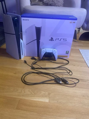 Sony PlayStation 5 med handkontroll - Säljer en PlayStation 5 konsol från Sony i vit och svart design. Medföljer en DualSense handkontroll, originalkartong samt kablar för ström och anslutning. Konsolen har en futuristisk look med slimmad form och diskret LED-belysning.