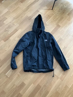 Svart regnjacka från The North Face - Snygg svart regnjacka från The North Face med huva och justerbar dragsko i nederkant. Jackan har dragkedja framtill, elastiska muddar vid ärmarna och logga på bröstet och ryggen. Perfekt för regniga dagar och enkel att packa ner i väskan.