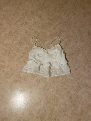 Vit broderad linnetopp med volang - Söt croppd linnetopp med tunna axelband och volangkant. Toppen har broderade blommor och är gjord i ett lätt material som passar perfekt till sommaren. Ger en romantisk och fräsch vibe till din outfit.