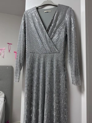 Glittrig silver långklänning omlott - Snygg långklänning i silver med glittrigt mönster och omlott framtill. Klänningen har v-ringning och långa ärmar, perfekt för dig som vill sticka ut. Materialet känns mjukt och följsamt, och den har en elegant siluett som passar till speciella tillfällen.