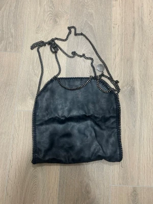 Svart Stella McCartney Falabella väska - Cool svart handväska från Stella McCartney, modell Falabella. Väskan har kedjedetaljer längs kanterna och axelrem i metall. Tillverkad i veganskt läder med en mjuk finish och fyrkantig form. Perfekt för dig som gillar trendiga accessoarer.