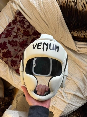 Vitt Venum huvudskydd - Säljer ett vitt huvudskydd från Venum, perfekt för boxning eller MMA. Skyddet har svart vaddering på insidan och justerbara band upptill för bra passform. Tillverkat i slitstarkt syntetmaterial och ger bra täckning för panna, kinder och bakhuvud. Endast använd en gång och nypris är runt 1000kr. Säljer även en svart vid intresse.