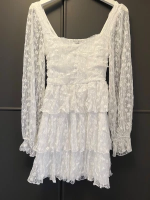 Vit spetsklänning med volanger - Romantisk vit kortklänning i spets med långa, transparenta ärmar och flera lager volanger. Klänningen har en fyrkantig ringning och är perfekt för somriga tillfällen eller student. Materialet är lätt och luftigt, vilket ger en feminin och elegant look.