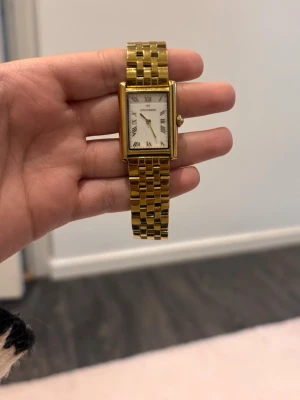 Guldfärgad klocka från Mockberg - Stilren klocka från Mockberg med fyrkantig urtavla i vitt och romerska siffror. Armbandet är guldfärgat och har en klassisk länkdesign. Perfekt accessoar för dig som gillar tidlös och elegant stil.