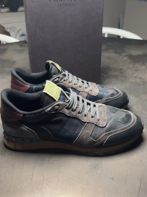Valentino Garavani camo sneakers - Snygga sneakers från Valentino Garavani med camomönster i svart, grått och olivgrönt. Skorna har detaljer i mocka och läder, samt nitar på hälen och en kontrasterande vinröd panel bak. Klassisk låg modell med grå snörning och gul logotyp på plösen.