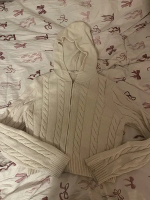 Beige stickad hoodie med dragkedja - Mysig beige stickad hoodie med dragkedja framtill och klassiskt flätmönster. Tröjan har långa ärmar, ribbade muddar och en stor huva. Perfekt för lager-på-lager och chill dagar. Materialet känns mjukt och värmande.