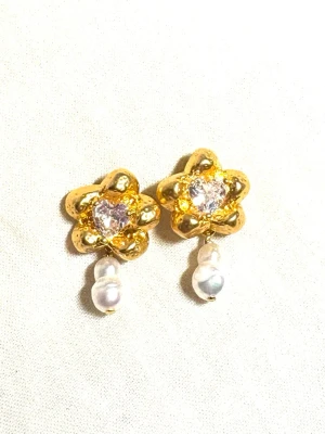 Guldiga blomörhängen med pärlor - Unika örhängen i guldfärgad metall formade som blommor med glittrande stenar formade som hjärtan i mitten och vita pärlor som hänger ned. Perfekta för dig som vill ha något som sticker ut och känns lyxigt. Säljs som par.