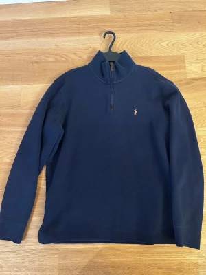 Half-zip tröja Polo Ralph Lauren - Klassisk marinblå tröja från Polo Ralph Lauren med half-zip och hög krage. Tröjan har det ikoniska broderade logotypen på bröstet och är tillverkad i mjukt bomullsmaterial. Perfekt för en stilren och avslappnad look.