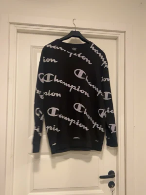 Svart Champion sweatshirt med logga - Säljer en svart sweatshirt från Champion med vit logotyp i stort tryck över hela tröjan. Tröjan har rund halsringning och långa ärmar. Perfekt för dig som gillar streetwear och vill ha en snygg och bekväm tröja.