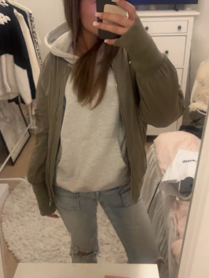 Olivgrön bomberjacka med fickdetaljer - Snygg olivgrön bomberjacka med dragkedja framtill och ficka med dragkedja på ärmen. Klassisk bombermodell med ribbade muddar vid ärmslut och nederkant. Perfekt att slänga över en hoodie för en avslappnad streetstyle-look.