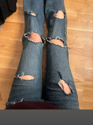 Blå slitna bootcut jeans - Säljer ett par blå bootcut jeans med flera stora slitningar och hål på framsidan. Jeansen har klassiska bakfickor med gul söm och är tillverkade i denim. Perfekta för en avslappnad och trendig look. (Köpta från Vinted)