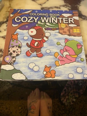 Cozy Winter målarbok - En färgglad målarbok med vintertema, fylld med söta djurfigurer som leker i snön. Omslaget har illustrationer av en hund, katt och kanin i vinterkläder framför ett hus. Perfekt för dig som gillar kreativa aktiviteter och gulliga motiv.