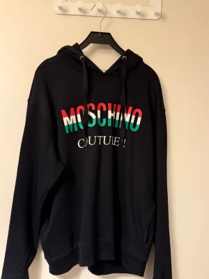 Svart Moschino hoodie med tryck - Snygg svart hoodie från Moschino med färgglatt tryck i rött, grönt och vitt på bröstet där det står 'MOSCHINO COUTURE!'. Hoodien har huva med svarta dragsnören och ribbade muddar vid ärmslut och nederkant. Perfekt för en trendig streetwear-look.