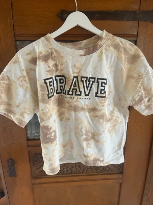 Beige tie-dye t-shirt från Lindex - Cool t-shirt från Lindex med beige och vita tie-dye mönster och texten 'BRAVE' framtill. T-shirten har korta ärmar och en ribbad rund halsringning. Perfekt för en avslappnad och trendig stil.