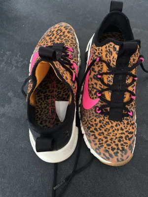 Nike sneakers leopardmönster & rosa - Unika Nike sneakers med leopardmönstrad ovandel, svarta detaljer och starkt rosa swoosh och snörhål. Skorna har vit och brun sula med rosa inslag undertill. Materialet är textil och syntet, och modellen är låg med svart hälflik och svarta skosnören.