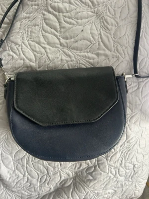 Svart axelväska i konstläder - Snygg svart axelväska i konstläder med justerbar rem och lock med kantig design. Väskan har en stilren look och passar perfekt till alla outfits. Enkel att bära över axeln och rymmer det viktigaste du behöver ha med dig. Innetygrt är lite slitet