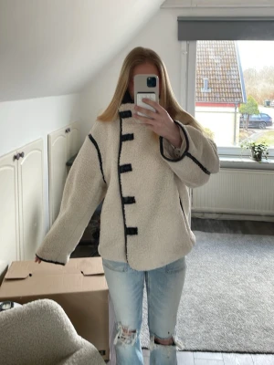Beige teddyjacka med svarta detaljer - Säljer en oversized beige teddyjacka med svarta kantdetaljer och knäppning framtill. Jackan har breda ärmar och en mysig, fluffig känsla. Perfekt för kalla dagar och ger en trendig look.
