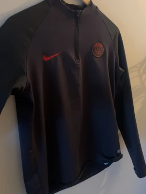 Svart PSG Nike Dri-FIT träningströja - Tröjan har halv dragkedja framtill och är tillverkad i Dri-FIT-material som andas. Perfekt för fotbollsträning eller gymmet. Den är Xl i barn storlek den passar för XS och S.