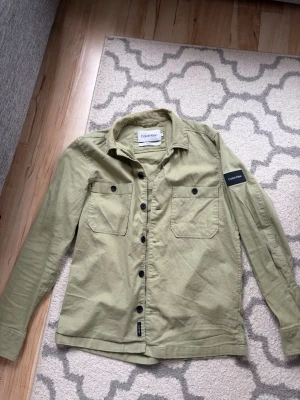 Beige overshirt från Calvin Klein - Stilren beige overshirt från Calvin Klein med två bröstfickor och svarta knappar. Overshirten har en klassisk krage och en svart Calvin Klein-patch på ärmen. Tillverkad i ett mjukt bomullstyg som ger en avslappnad look.