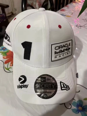 Vit Red Bull Racing keps - Säljer en vit Red Bull Racing keps från New Era 9FORTY-serien. Kepsen har broderad Oracle Red Bull Racing-logga framtill och färgglad brodyr med sol, vågor och moln på sidan. Justerbar passform och röda detaljer på ovansidan. Perfekt för motorsportfans!