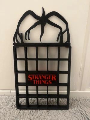 Stranger Things hylla  - Säljer en svart hylla med Stranger Things-tema, tillverkad i plast. Hyllan har ett rutnät med plats för småsaker och en cool siluett av en Demogorgon på toppen. Texten 'Stranger Things' i rött finns i mitten. Perfekt för fans av serien!