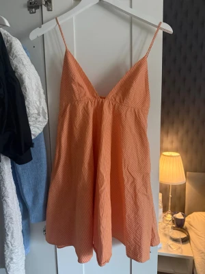 Randig orange sommarklänning - Säljer en luftig orange klänning med tunna vita ränder och smala axelband. Klänningen har en v-ringad hals och ett avslappnat, flowy fall. Perfekt för varma dagar och enkel att styla med sneakers eller sandaler.