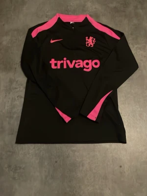 Svart Nike tränings-tröja Chelsea - Snygg svart långärmad tränings-tröja från Nike med rosa detaljer och Chelsea FC-logga på bröstet. Tröjan har rosa Nike-logga och 'trivago' tryckt framtill. Perfekt för fotbollsträning eller gymmet. Tillverkad i funktionsmaterial som andas.