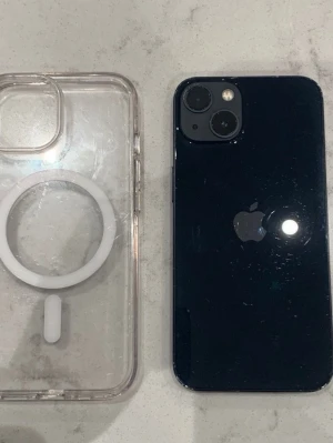 iPhone 13 svart med MagSafe-skal - Säljer en svart iPhone 13 från Apple med stilren design och dubbla kameror på baksidan. Telefonen har en slimmad form och kommer med ett genomskinligt MagSafe-kompatibelt skal i plast. Perfekt för dig som vill ha en modern och snygg smartphone. Har en liten spricka.