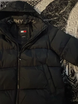 Svart pufferjacka från Tommy Jeans - Snygg svart pufferjacka från Tommy Jeans med huva och dragkedja framtill. Jackan har broderad logga på insidan av kragen och klassisk quiltad design. Perfekt för kalla dagar och riktigt skön att bära. Passar dig som vill ha en stilren och varm vinterjacka.