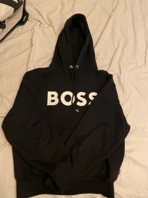 Svart oversized hoodie från BOSS - Snygg svart hoodie från BOSS med oversized fit och stor vit logga på bröstet. Hoodien har dragsko i huvan och ribbade muddar vid ärmar och nederkant. Perfekt för en avslappnad och trendig stil.