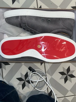 Gråa Christian Louboutin sneakers storlek 42 -  Pris kan diskuteras!! Snygga gråa sneakers från Christian Louboutin i mocka med klassisk röd sula och vita kanter. Lågt skaft och minimalistisk design med ton-i-ton snörning. Perfekta för dig som vill ha något exklusivt och stilrent. 