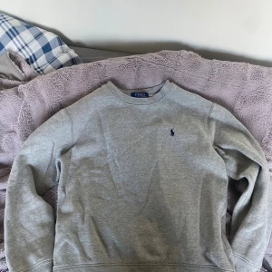 Grå sweatshirt från Polo Ralph Lauren - Grå sweatshirt från Polo Ralph Lauren i storlek M. Klassisk crewneck med broderad marinblå logga på bröstet. Tröjan har långa ärmar och är gjord i mjuk bomullsmix, perfekt för chill dagar. Enkel och stilren design som funkar till jeans eller joggers.