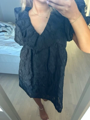 Svart kort klänning med volang - Snygg svart kort klänning med volangdetaljer och v-ringning. Klänningen har puffiga korta ärmar och en lätt strukturerad yta som ger en cool look. Perfekt för dig som vill sticka ut med en trendig och bekväm outfit. Använd en gång💕