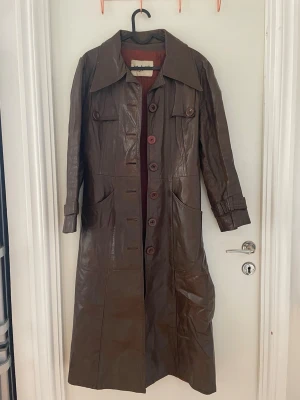 Brun lång skinnkappa med bälte - Snygg brun lång läderkappa med stora knappar och bälte i midjan. Kappan har bred krage, fickor framtill och dekorativa detaljer på ärmar och rygg. Perfekt för dig som vill ha en retro och cool look. Materialet är äkta läder.