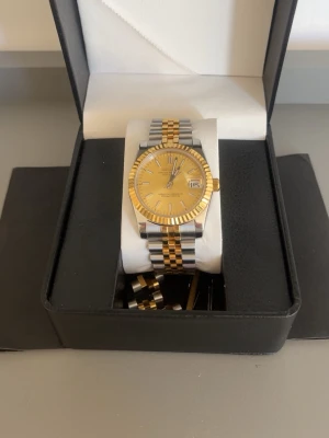 SEIKO MOD  Datejust guld och silver klocka - Säljer en snygg SEIKO MOD Datejust klocka med tvåfärgad länk i guld och silver. Urtavlan är guld med datumvisning och klassiska index. Länken har ett randigt mönster och är tillverkad i metall. Perfekt för dig som vill ha en stilren och lyxig accessoar. ALLA LÄNKAR FINNS MED 36mm. Kan gå ner i pris vid snabb affär. Har kvitto